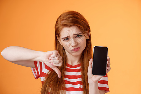 质量很差 心烦意乱的红发女顾客表情失望 显示智能手机空白显示 大拇指向下判断 不喜欢应用程序 假笑沮丧 站在心烦意乱的橙色背景电图片
