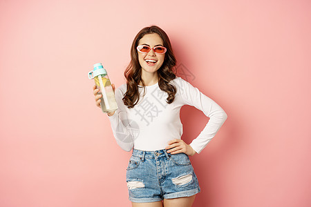 穿春装的时髦女孩 穿着太阳眼镜 拿着柠檬水瓶 健康饮酒 笑笑和笑 站在粉红背景上女性喜悦乐趣互联网魅力黑发广告微笑工作室青年图片