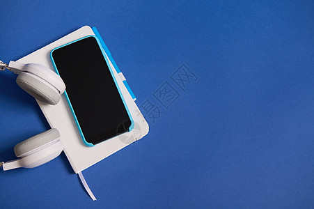 耳机平躺 蓝色外壳的智能手机放在白色日记组织者身上 与蓝色背景隔离 带有复制空间 白色和蓝色办公工具的顶视图图片