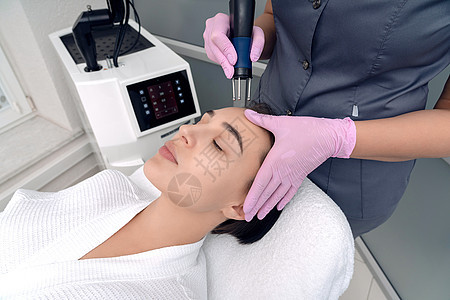 美容 美丽的女人在 Spa 诊所接受治疗师的刺激性电动面部护理 微电流治疗期间年轻女性面部特写图片