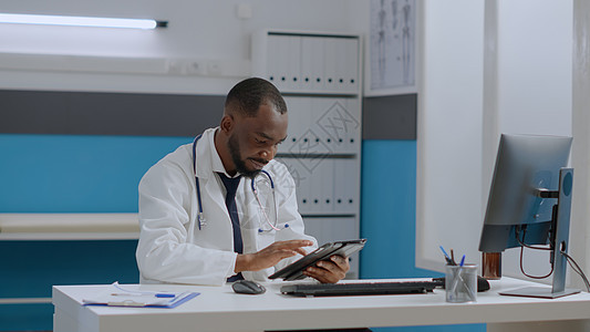 坐在医院办公室服务台的非洲-美国治疗师医生持有平板电脑(Patst计算机)图片