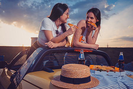 两个年轻女孩在吃比萨饼 大笑 用黄色汽车装扮薯条 帽子和汽水 放在箱子的玻璃瓶里 快餐图片