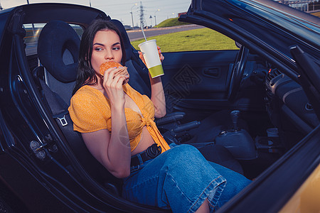 蓝色牛仔裤和橙色顶部的模型是吃汉堡包 坐在黄色轿车上时用纸杯装饮料 快餐 复制空间图片