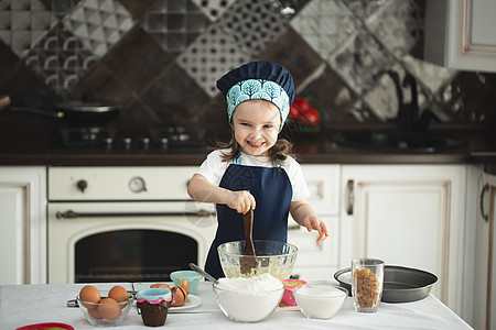 一个穿着围裙的可爱小女孩 和主厨的帽子 用木制毛片搅钱 看着摄影机 一边笑着做饭乐趣情感假期糕点面包师舌头面包烹饪手工女性图片