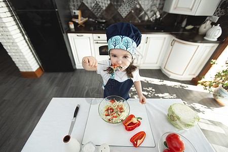 穿着围裙和厨帽的小孩 在厨房吃蔬菜沙拉 看着摄像机 (笑声)图片