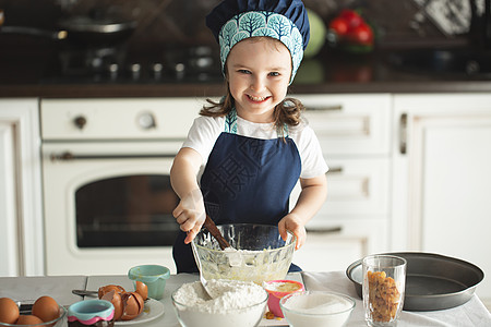 面粉蛋糕一个穿着围裙的可爱小女孩 和主厨的帽子 用木制毛片搅钱 看着摄影机 一边笑着做饭面粉桌子厨师童年滚动女性厨房食谱烹饪情感背景