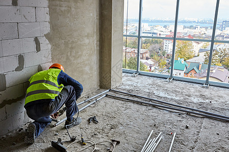 身着工作服和安全帽的建筑工人正在正在建设中的带有全景窗户的建筑物的公寓内安装塑料管道 高层住宅图片