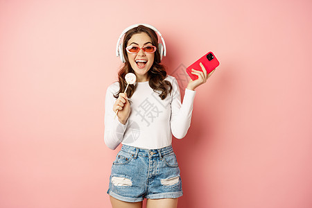 暑期女孩笑 吃润滑油和耳机听音乐 用智能手机在粉红背景下跳舞广告成人头发女性标识发型黑发乐趣冒充快乐图片