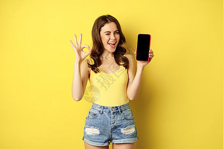 漂亮的黑发女人 展示手机屏幕 智能手机应用程序界面和好的标志 推荐网站 商店 站在黄色背景下女性魅力细胞女孩工作室成人电话技术冒图片