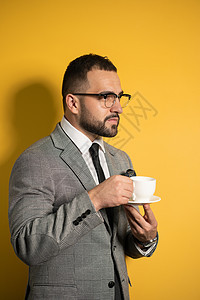戴着眼镜 戴着灰色西装 拿着一杯咖啡 留着胡须的英俊年轻商务人士 一边喝着咖啡 一边站在一边 与黄色背景隔绝图片