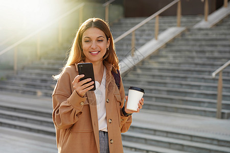 带着智能手机微笑的年轻商业女青年在早晨拿起咖啡 背景上楼梯模糊不清 而且早上带走咖啡景观城市技术女孩拉丁拿铁学生脚步街道外套图片