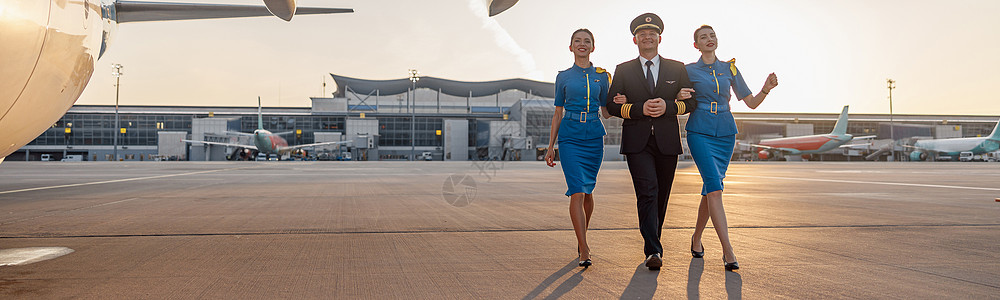 兴奋的男性飞行员和两名身穿蓝制服的女性空服女乘务员在日落时候终点站飞机前行走 一整拍全镜头男人喷射服务员航空公司团队航班全体旅游图片