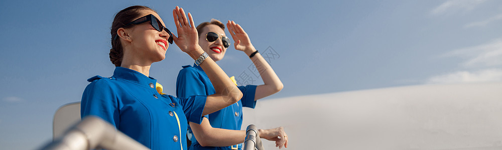 两个穿蓝色制服的优雅空姐和太阳镜 双手用眼罩着眼睛 望远相望 站在一起在空中的肖像冒充人员服务员水平职业乘客楼梯飞机旅行航班图片