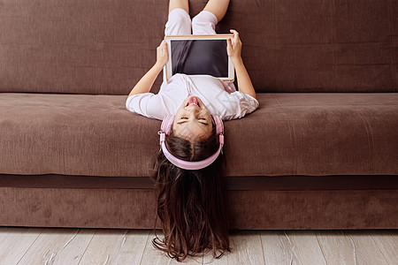 一个穿着粉红家庭衣物和粉红色耳机的有趣的小女孩 躺在沙发上 带数码平板电脑图片