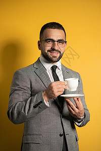 英俊的大胡子年轻人戴着眼镜 穿着正装 手里拿着一杯咖啡 背景是黄色的 商业 热饮和人的概念胡须管理人员职业男人经理早餐工作室杯子图片