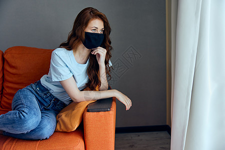 穿着面罩的漂亮女人坐在家中橙色沙发上 呆在家里感染疾病药品暴发流感恐慌卫生安全成人窗户图片