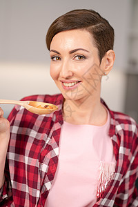 穿着红色格子衬衫和在现代厨房做短发烹饪的短发汤的近身家庭主妇 微笑着的妇女测试她的菜盘图片
