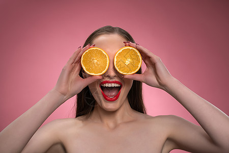 有趣的美女模特年轻女子拿着两片多汁的橙片作为她的眼睛或眼镜 迷人 快乐 风趣的女士 红唇 长发 粉红色背景中突显图片