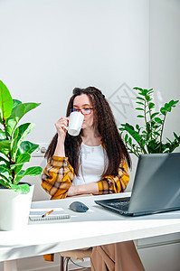 戴眼镜的漂亮女人在工作场所休息时喝咖啡 妇女在家里工作 使用笔记本电脑图片