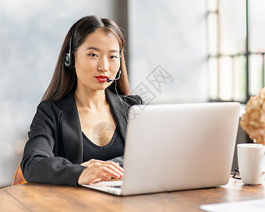在耳机上快乐的亚洲女商务人士通过电话会议和视频聊天来发言商业人士笔记本接待员监视器办公室电脑热线秘书服务图片