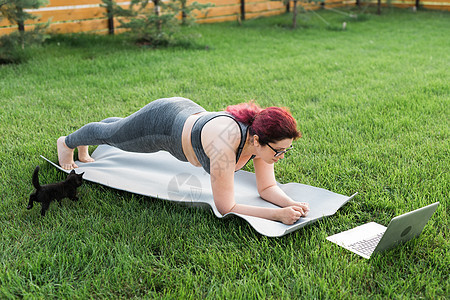 身穿运动上衣和紧身裤的年轻加体型女性站在瑜伽垫上的木板上 在院子里的绿草上消磨时光 黑猫在她身边走来走去 健康和健身概念女孩肚子图片