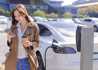 微笑的年轻女性站在电动车附近的城市停车场 从小城市站为汽车电池充电 喝咖啡和使用智能手机收费力量司机驾驶活力车辆女士杯子服务商业图片