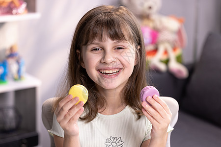 一个小女孩在彩色家庭背景下用蛋白杏仁饼干遮住眼睛的快乐笑脸 快乐 十几岁的女孩玩甜点马卡龙 把饼干像眼镜一样放在眼睛周围图片