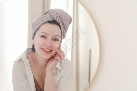 头戴毛巾的年轻女子看着镜子 在脸皮上涂奶油皮肤健康粉刺口罩女士微笑情感水分清洁擦洗图片