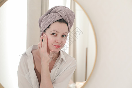 头戴毛巾的年轻女子看着镜子 在脸皮上涂奶油情感粉刺女性温泉健康皮肤化妆品福利女士口罩图片