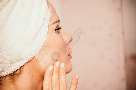 年轻漂亮的女人在洗完澡后使用保湿面膜 头戴毛巾的漂亮迷人女孩站在家庭浴室的镜子前 日常卫生和皮肤护理产品粉刺治疗美容女士皱纹预防图片