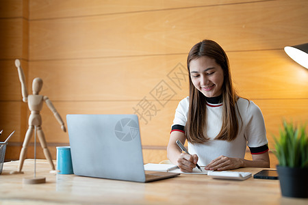 微笑的女学生穿着做笔记与老师一起在线学习 快乐的年轻女人学习语言听讲座看网络研讨会写笔记看家里的笔记本电脑 远程教育图片