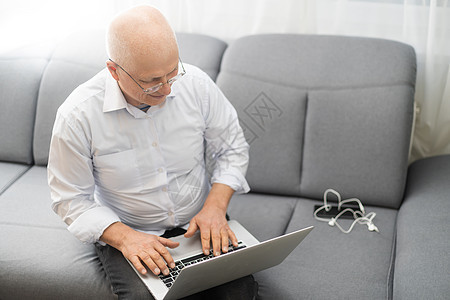 穿白衬衫和黑裤子的老年秃头男子在客厅里修眼镜和用笔记本电脑工作工具成人社交男性职业技术冲浪互联网退休媒体图片