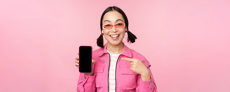 热情的亚洲女性穿着时尚的衣服 戴着太阳镜 手指指着手机屏幕 展示智能手机应用程序 站在粉红色背景上细胞购物女孩办公室界面标识女朋图片