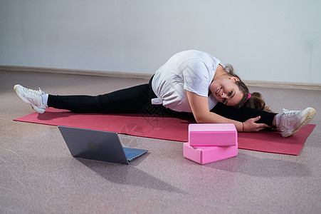 年轻的白人胖女人在运动垫上弯腰 在笔记本电脑上观看训练视频 微胖妹利用视频通讯远程拉开分叉学生肌肉灵活性女士锻炼动机重量运动服地图片