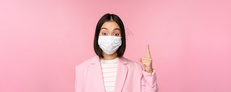 穿着医疗面罩和西装的亚洲女商务人士贴近肖像 举起手来 展示广告 头条横旗 站在粉红背景上标识人士企业家健康商务经理女士工作售货员背景图片