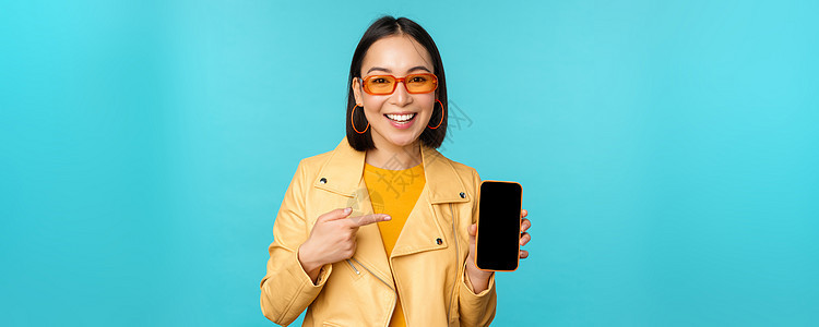 年轻时尚的亚洲女模特戴着时髦太阳镜 展示手机屏幕 在智能手机界面上应用 站在蓝色背景上成人情绪女孩微笑商业企业家工作室促销办公室图片