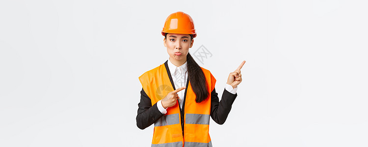建筑 施工和工业概念 心烦意乱的亚洲女经理 安全头盔工程师噘嘴 抱怨坏事 指着右上角装修安全企业快乐反光头盔电子商务女性工作房地图片