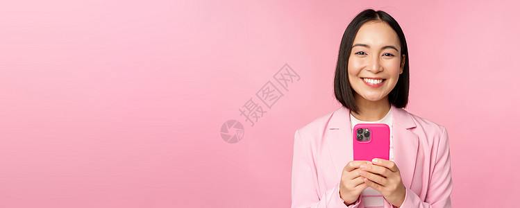 使用智能手机 移动电话应用程序 站在粉红背景之上的Asian公司老板 微笑着笑容的商业妇女的肖像办公室购物广告售货员电话互联网职图片