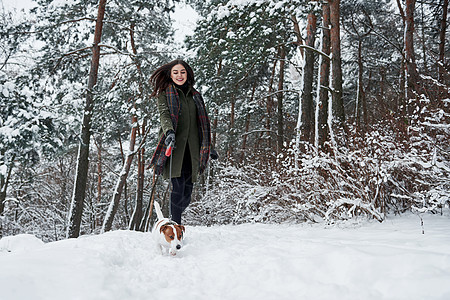 宠物主人平凡的一天 穿着暖和衣服的女人在白雪皑皑的森林里遛狗 正视图图片
