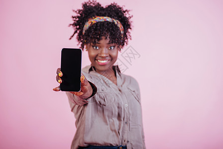 微笑并感到快乐 手里拿着黑色手机 工作室里粉红色背景中穿着休闲服的迷人非洲裔美国女性图片