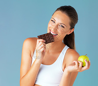 我的饮食为偶尔的放纵腾出了空间 在蓝色背景下 一位健康的年轻女子拿着苹果咬了一口巧克力的摄影棚照片图片