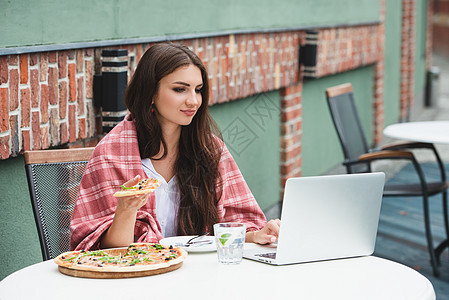 年轻的自由职业妇女使用笔记本电脑 坐在咖啡桌边吃披萨闲暇教育女性女孩笔记桌子女士商业互联网学习图片