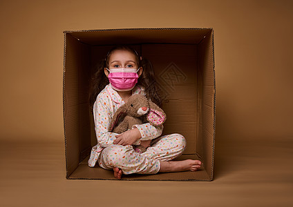 穿着睡衣 穿着粉红色医疗保护面罩的白种女婴拥抱她 坐在纸板盒子里 看着照相机 在米底背景上与复制广告空间隔绝图片