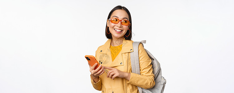 时尚的年轻女性女游客 带背背包和智能手机旅行者在镜头下微笑 以白色背景装扮太阳镜购物工作室办公室电话成人广告促销黑发大学图片