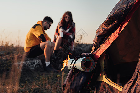 以垫子和帐篷为焦点的背包 背景是日落时坐在篝火旁的两个年轻旅行者男人和女人 旅行概念图片