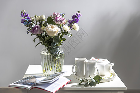 玻璃花瓶中的 紫色和白色玫瑰 小茶玫瑰 和蓝色鸢尾花束放在白色咖啡桌上 还有两个高脚茶杯 灰墙图片