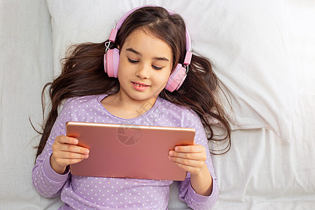 戴着粉色耳机的漂亮黑发女孩躺在床上 看着粉色数字平板电脑图片
