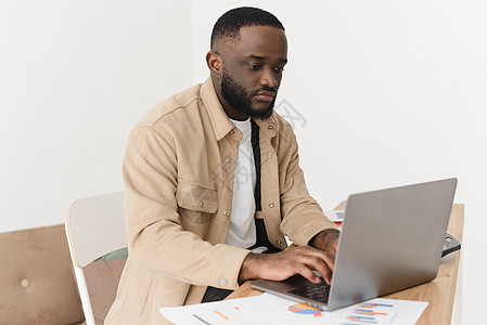 专注的年轻美国黑人男子计算开支并在笔记本电脑上记录 纸质银行账户在桌子上 男模特使用电子银行计算机应用程序管理每月家庭预算 计划图片