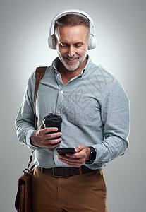 他是一个积极进取的人 总是为新的一天做好准备 摄影棚拍摄的是一个成熟男子戴着耳机 在灰色背景下拿着袋子和咖啡使用手机背景图片