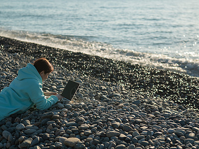 白种女人躺在卵石沙滩上的肚子上 打笔记本电脑 远距离工作自由职业者网络航班太阳自由旅行女性娱乐海岸码头键盘图片